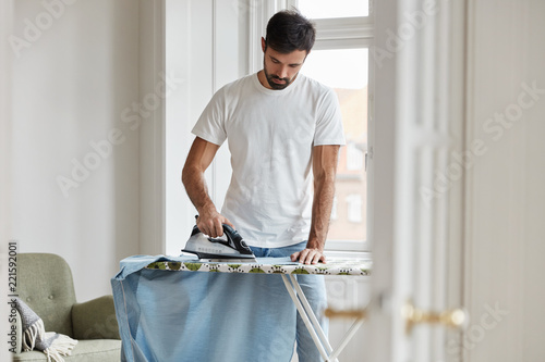Photo Hard working young Caucasian bearded husband irones clothes on ironing board, does domestic duties while wife is away, stands in living room, being busy with work about house
