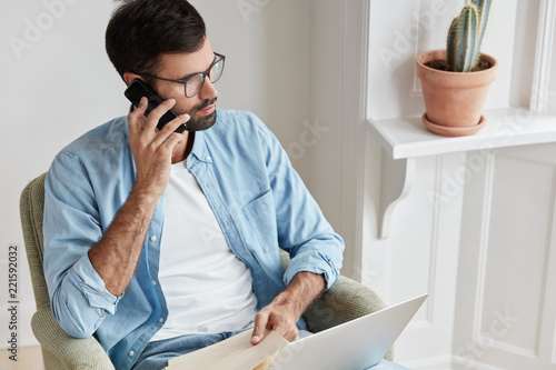 Фотография Shot of IT developer calls to costumer service, makes transactions, checks balance, uses modern cell phone and laptop computer for communication and work, does distance job