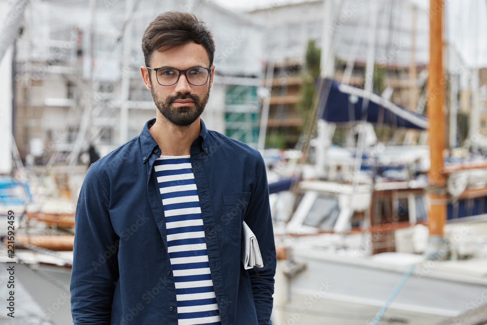 Outdoor shot of bearded young sailor dressed in fashionable clothes, has rest after arrival, poses against sailboats blurred background, carries newspaper under arm, breathes fresh marine air Outdoor shot of bearded young sailor dressed in fashionable clothes, has rest after arrival, poses against sailboats blurred background, carries newspaper under arm, breathes fresh marine air