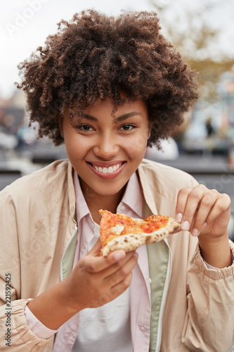 Vertical shot of beautiful Afro American girl has delicious pizza after hard working day, dressed in casual jacket, visits outdoor cafeteria, has broad shining smile. People and lunch concept