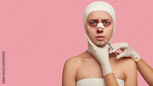 Mentoplasty and lip plumping concept. Young girl recieves cosmetic injection for lip augmentation, has bandage on nose after rhinoplasty, good medical treatment. Surgeon holds syringe near face photo