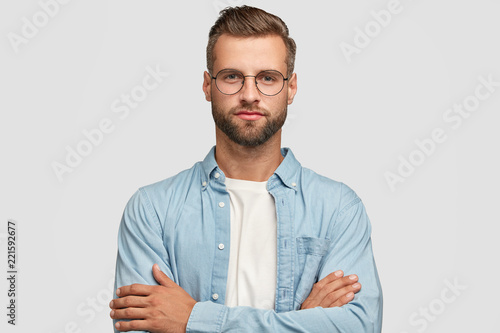 Horizontal shot of handsome self confident businessman or designer, stands crossed hands against white background, wears casual shirt, poses against white background. People and lifestyle concept