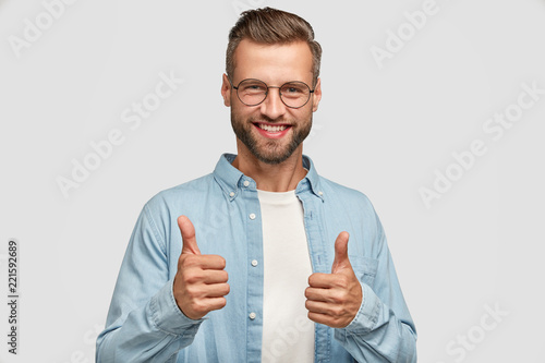 Tablou canvas Handsome young Caucasian bearded man does okay symbol, keeps thumb raised, approves good idea of companion, has cheerful expression, stands alone agaisnt white background
