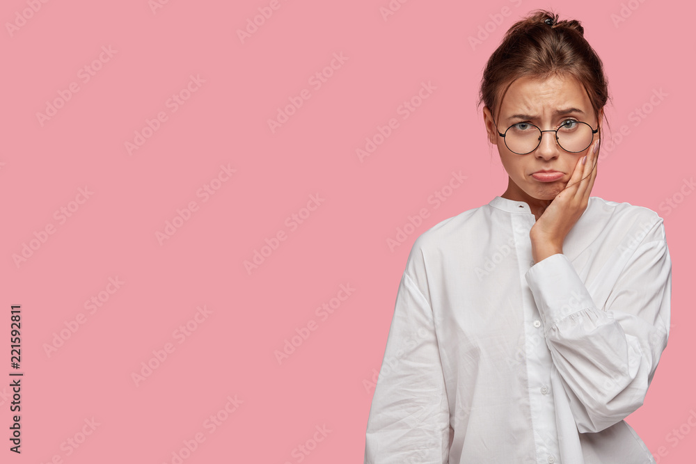 Photo of beautiful offended young woman has toothache, purses lower lip, dressed casually, wears round spectacles, isolated over pink background with copy space for your advertising content.