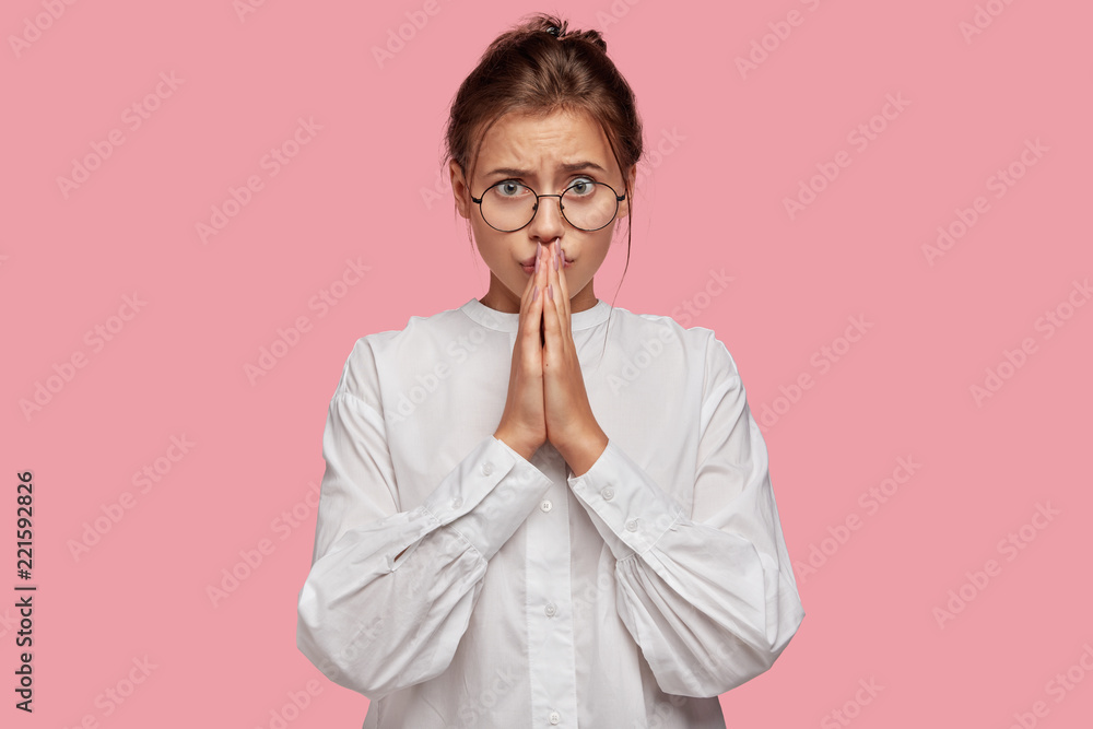 Horizontal shot of pretty nervous student prays before passing exam, has embarrassed expression, poses against pink studio wall. Woman believes in good luck and wellness keeps palms in praying gesture Horizontal shot of pretty nervous student prays before passing exam, has embarrassed expression, poses against pink studio wall. Woman believes in good luck and wellness keeps palms in praying gesture