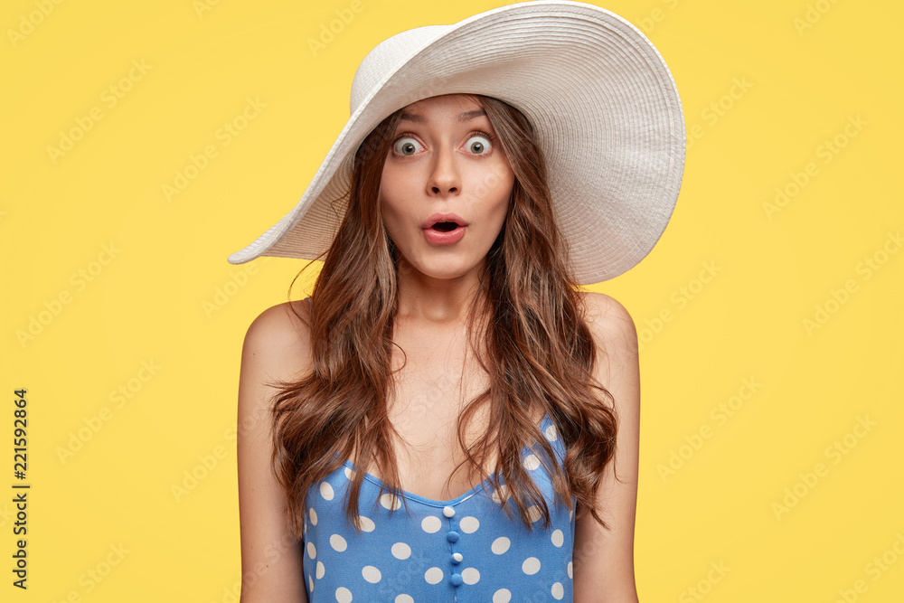 Emotive surprised fashionable woman in summer hat, stares with bugged eyes, opens mouth from surprisement, dressed in polka dot clothes, stands against yellow background. Surprisement concept - obrazy, fototapety, plakaty 