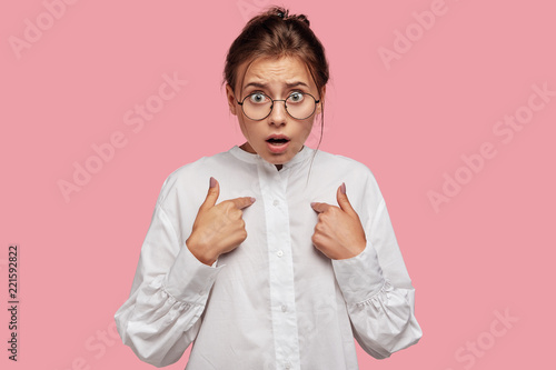 Photo of indignant beautiful Caucasian woman points at herself with discontent expression, being stupefied to have many duties at work, dressed in white shirt, poses against pink background. photo