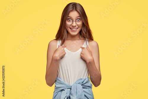 Studio shot of indignant woman with pleasant appearance, points at herself as asks she really won prize, has cheerful expression, dressed in casual vest and shirt around waist, isolated on yellow