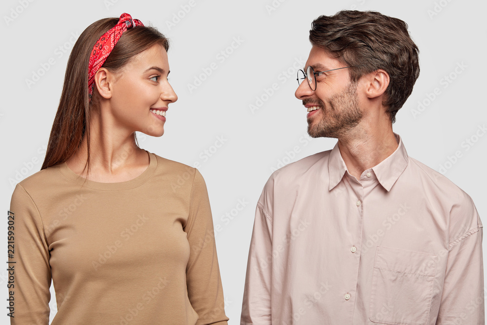 Pretty young woman and her boyfriend stand against white background, look at each other with positive expressions, have good friendly relationships. Groupmates meet together for making common task Pretty young woman and her boyfriend stand against white background, look at each other with positive expressions, have good friendly relationships. Groupmates meet together for making common task