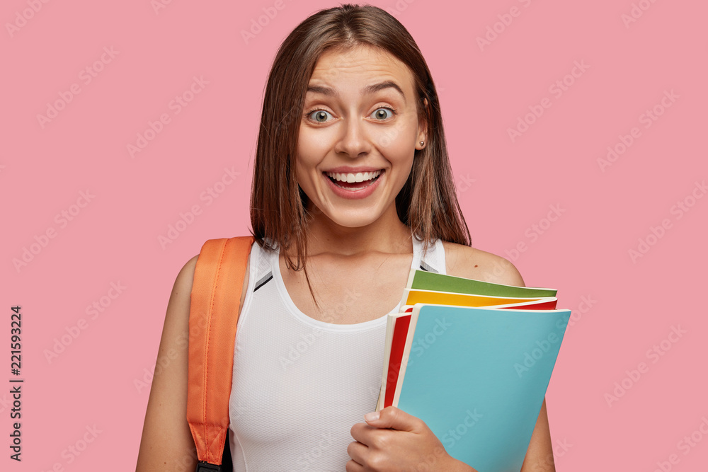 Photo of cheerful European girl with broad smile, dressed in casual white t shirt, prepares for learning material, carries rucksack, isolated over pink studio wall. Reader visits library. Indoor shot Photo of cheerful European girl with broad smile, dressed in casual white t shirt, prepares for learning material, carries rucksack, isolated over pink studio wall. Reader visits library. Indoor shot