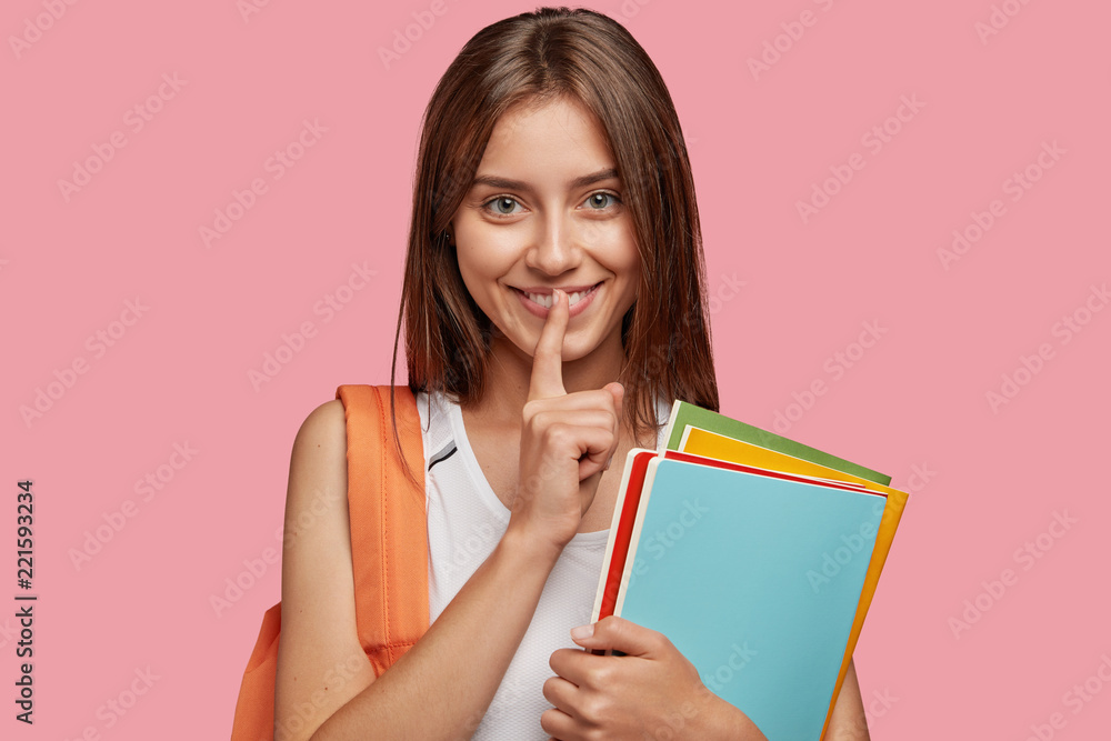 Education in high school. Pleased young Caucasian woman keeps fore finger on lips, makes secret gesture, holds folders for paper documents, gossips about boyfriends with true friend, stands indoor Education in high school. Pleased young Caucasian woman keeps fore finger on lips, makes secret gesture, holds folders for paper documents, gossips about boyfriends with true friend, stands indoor