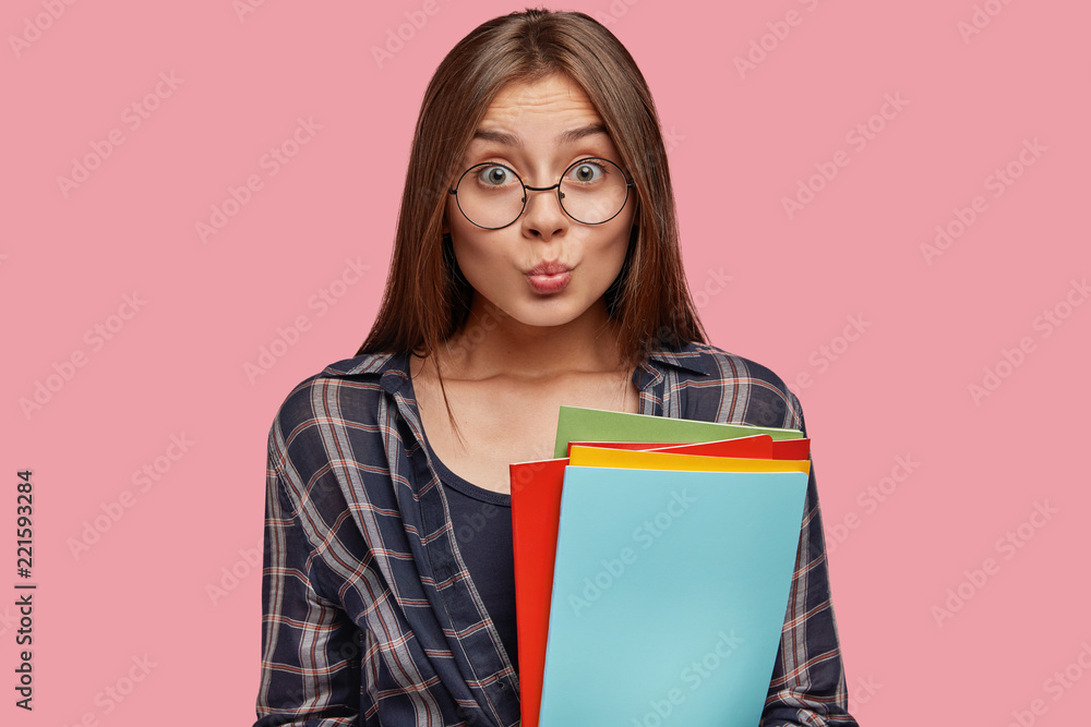 Attractive businesswoman holds business folders, makes grimace, dressed in checkered shirt, ready to prepare report, isolated over pink background. People, education and facial expressions concept Attractive businesswoman holds business folders, makes grimace, dressed in checkered shirt, ready to prepare report, isolated over pink background. People, education and facial expressions concept