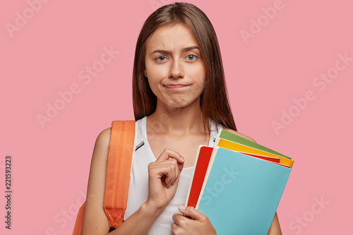 People, studying and university concept. Puzzled clever intern listens attentively lecture, holds colourful folders, carries rucksack, stands against pink background, purses lips in bewilderment.