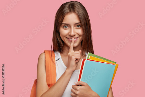 Education in high school. Pleased young Caucasian woman keeps fore finger on lips, makes secret gesture, holds folders for paper documents, gossips about boyfriends with true friend, stands indoor
