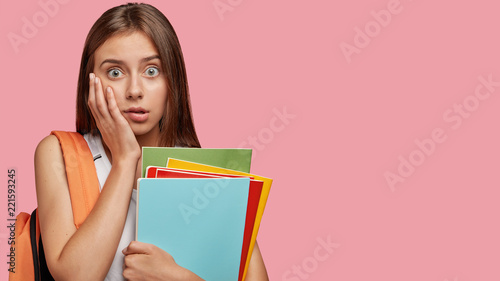 Studio shot of pretty woman with stunned expression, keeps hand on cheek, cant believe in failure, holds copybooks or textbooks, isolated over pink background with copy space for your advertisement