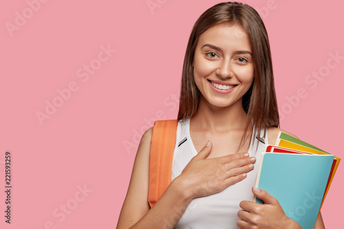 Friendly pleased thankful young hipster carries rucksack, holds colourful folder with papers, keeps hands on chest, has tender smile, isolated over pink background with copy space for your text