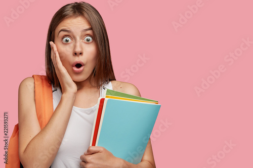 Horizontal shot of stupefied European woman keeps eyes widely opened, jaw dropped, terrified with schedule at college, carries books or textbooks, isolated over pink background with copy space