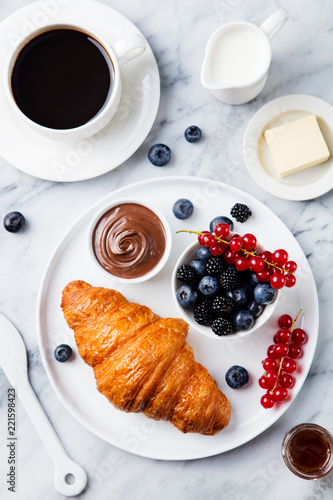 Croissant with fresh berries, chocolate spread and butter with cup of coffee on a marble background. Top view.