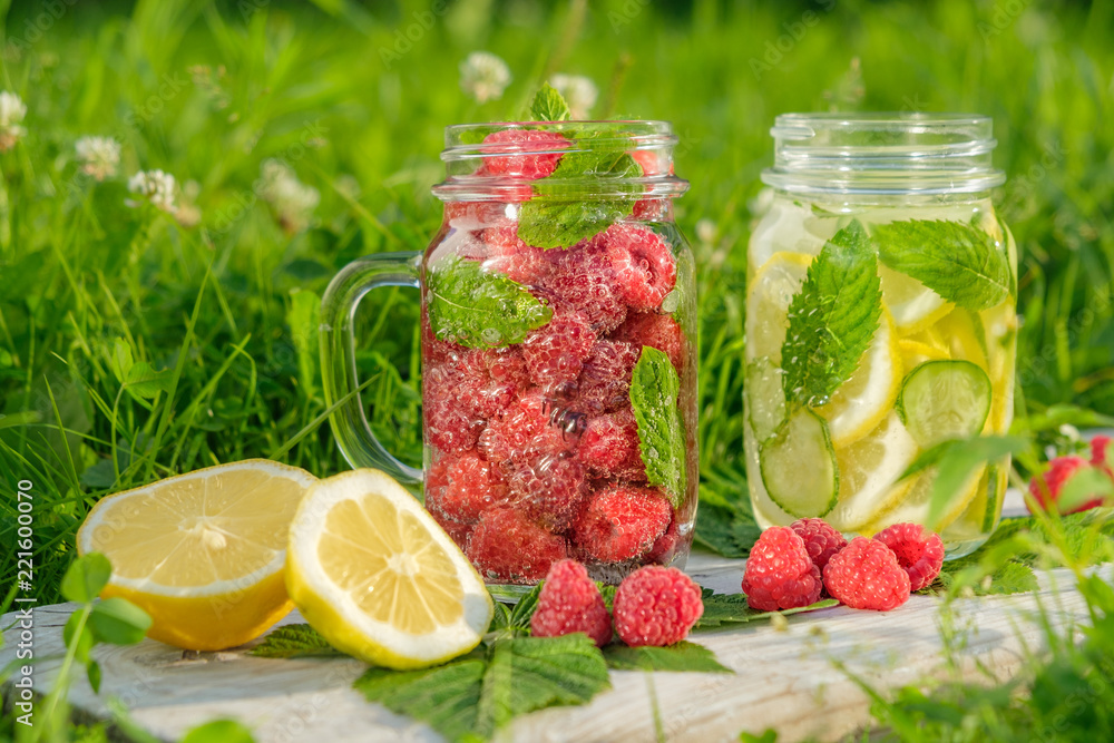 Lemonade with raspberries and lemonade with lemon, cucumber and mint in a hipster jar for jam on a background of green grass. Concept: a picnic with refreshing drinks Lemonade with raspberries and lemonade with lemon, cucumber and mint in a hipster jar for jam on a background of green grass. Concept: a picnic with refreshing drinks