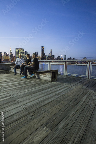 Three people sitting on bench at dusk at Gantry State Plaza Park on the East River waterfront in Long Island City, Queens, NY with Manhattan skyline in distance photo