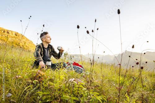 A young traveler sits on a hill in the background of a mountain and drinks tea from a thermos