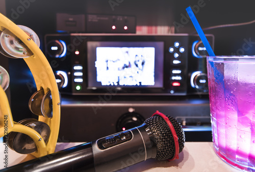 Black microphone in karaoke club, with remote controller, melon and strawberry soda drinks, yellow tambourine and screen for singing music on stage party.