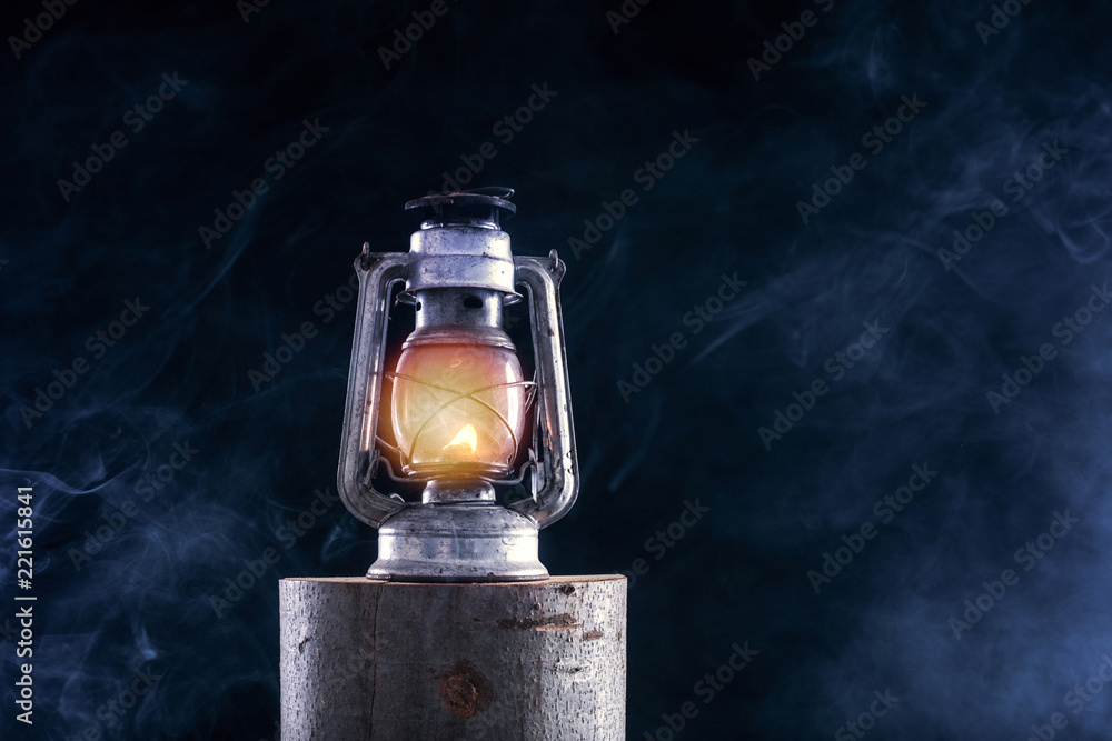 Old oil lamp burning at lumber log and foggy smoke dark night background. Rust metal lantern with corrosion. Horror and Halloween concept. Close up, selective focus