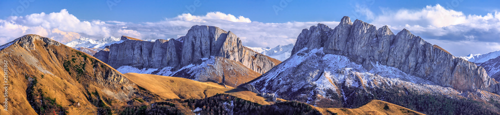 Beautiful scenic autumn landscape of majestic Acheshbok rocky mountain peaks called Devil's Gate covered with snow under blue sky with clouds. Wide panoramic mountain scenery in West Caucasus, Russia