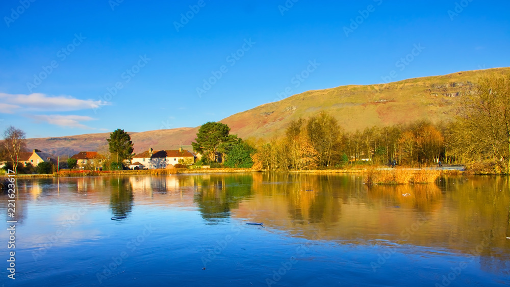 Reflections of trees and a clear blue sky on a rural village pond.