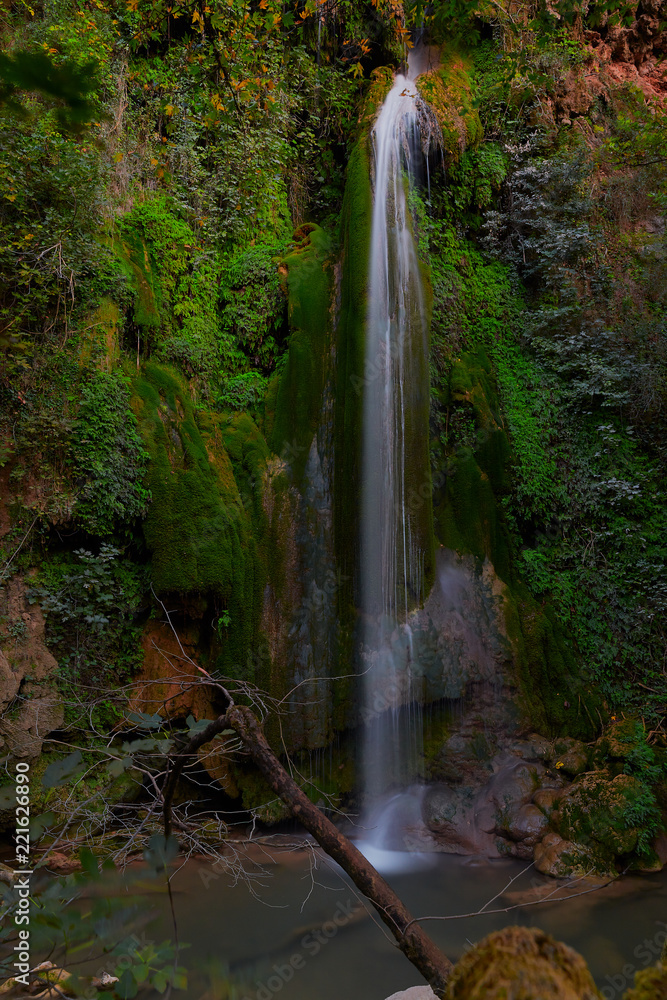 Fototapeta premium Blurred waterfall comes from inside the red rocks with Green trees and plants around