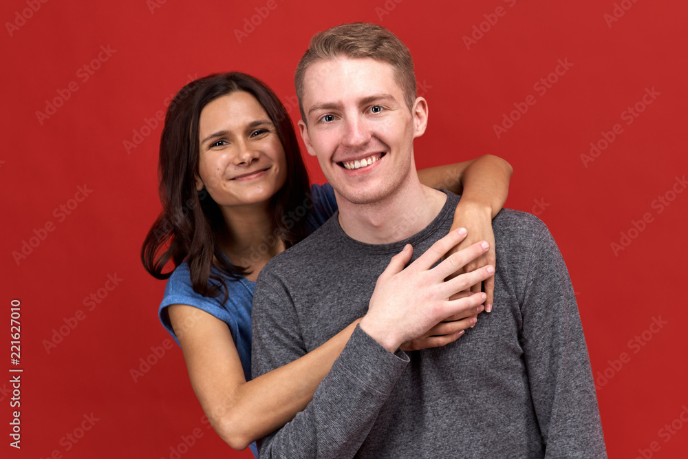 An attractive young guy smiles widely because his girlfriend hugs him. Portrait of a happy couple of friends, relationships and close feelings. Studio portrait on red background An attractive young guy smiles widely because his girlfriend hugs him. Portrait of a happy couple of friends, relationships and close feelings. Studio portrait on red background