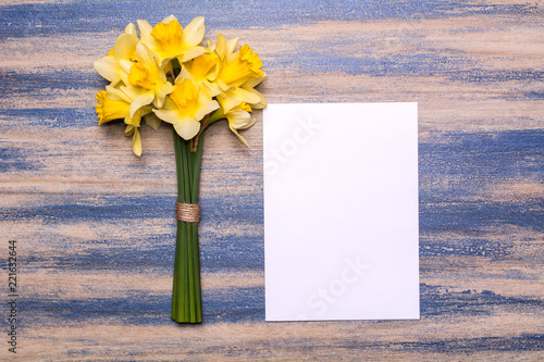 Fototapeta Naklejka Na Ścianę i Meble -  A bouquet of daffodils and a sheet of white paper on a wooden background. Spring yellow flowers. Paper with space for text. Blue peeling paint. Flat lay design, top view.