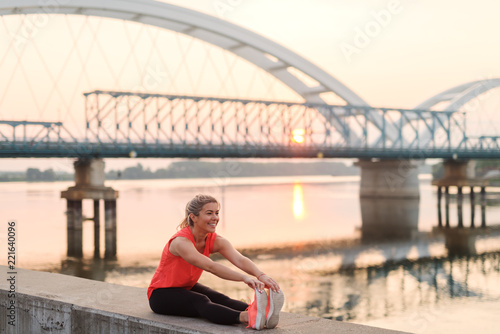 Beautiful sporty blond girl stretching her muscles by the river and enjoying quiet summer morning.