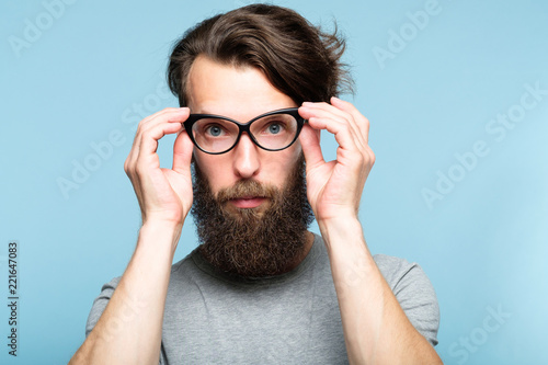 bearded hipster guy fixing his cat eye glasses. stylish modern fashionist. portrait of a geeky quirky eccentric man on blue background.