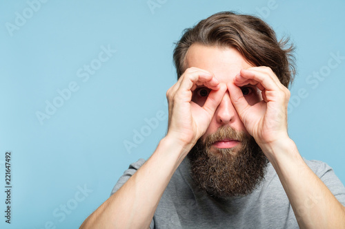 funny ludicrous joyful comic playful man pretending to look through binoculars made of hands. portrait of a young bearded guy on blue background. emotion facial expression concept photo