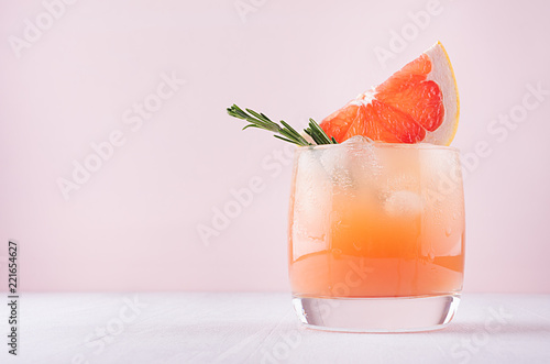 Cold fresh homemade grapefruit cocktail with ice, rosemary and pieces citrus on pastel pink background.