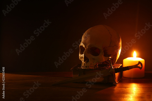 Human skull, old book, sword and burning candle over old wooden table and dark background.