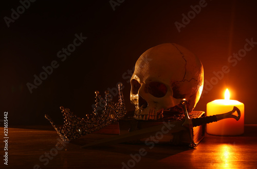 Human skull, old book, sword, crown and burning candle over old wooden table and dark background.