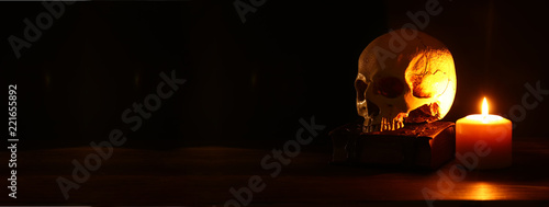 Human skull, old book and burning candle over old wooden table and dark background.