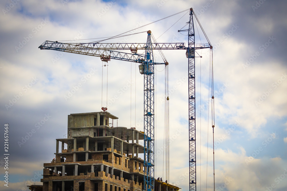Two building cranes in front of the building under construction against a sky with clouds