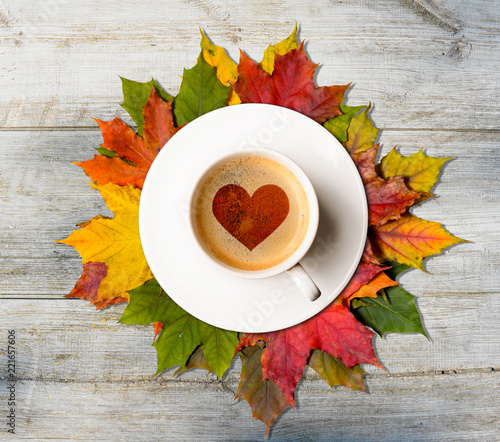 Fall in love. Coffee cup with heart symbol on autumn colorful leaves on wooden table, top view