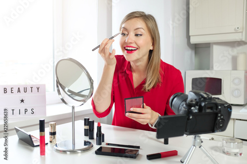 Stylish girl blogger in red blouse drawing on her eyebrows, looking at mirror, while recording make up video guide. Beauty tips