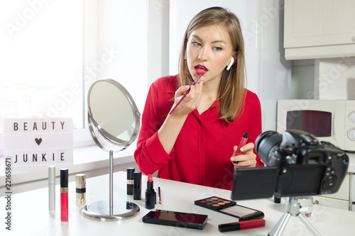 Female beauty blogger putting on red lipstick on her lips, looking at video camera, sitting at kitchen table at home