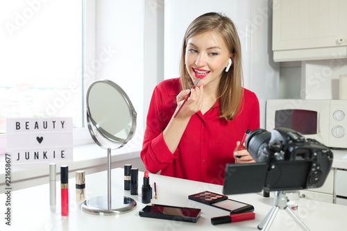 Portrait of pretty young female video blogger, wearing stylish red blouse, sitting and using brush to apply lipstick, looking at camera
