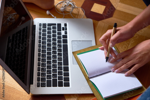 Top view close-up of legs and hands of young female sitting on the wooden floor with a laptop, pen, notebook and earphones. Lifestyle. Home office concept