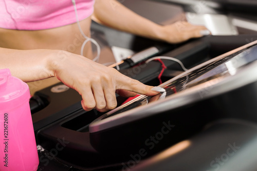 slim woman standing on the running track in the gym wearing earphones listening to music and setting the speed. Concept of cardio exercises and healthy way of life