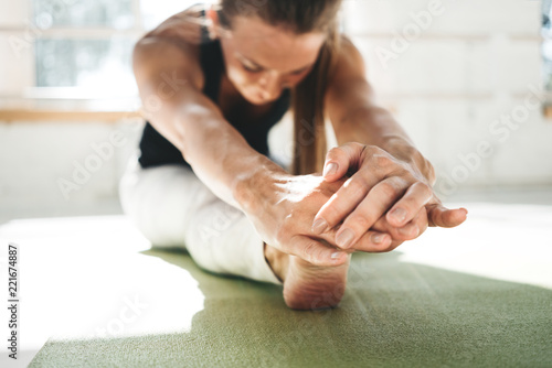 Female athlete doing stretching exercises listening in sunny sport gym in the morning. Fitness strong woman bending forward stretching her leg sitting on yoga mat photo