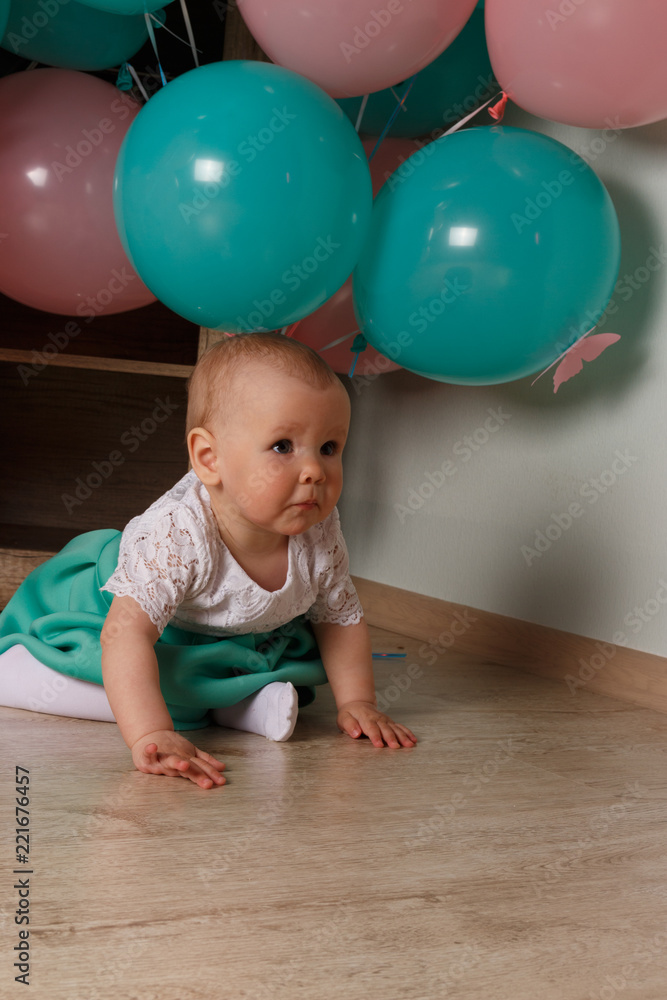 a small child, a girl, a baby, sits on the floor in a white and blue dress, against a background of blue and pink air, gel balls. the child is not happy, sad.
