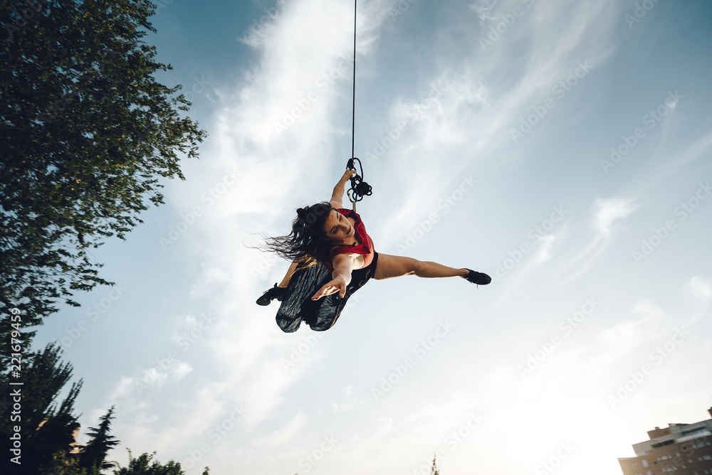 Sexy dancer performing aerial dance hanging on harness on urban scenery at sunset against blue sky Sexy dancer performing aerial dance hanging on harness on urban scenery at sunset against blue sky