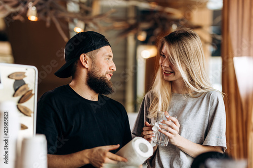 Two young smiling people,a blonde girl and man with beard.dressed in casual outfit, stand next to each other and look at each other in a cozy coffee shop.